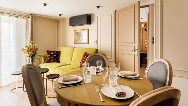 A dining area with a round table set for four, a sofa with cushions, a vase of flowers, and a television mounted on the wall. - Merveil - Luxury Suite - Marais - Turenne (Paris)