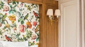A hotel room with a floral wallpaper, a bed with white linens, two decorative pillows, a wooden headboard, a wall-mounted light fixture, and a door with a curtain. - Merveil - Luxury Suite - Marais - Turenne (Paris)