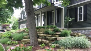 Una casa residencial con porche delantero, escalones de piedra y un jardín ajardinado con diversas plantas y arbustos.