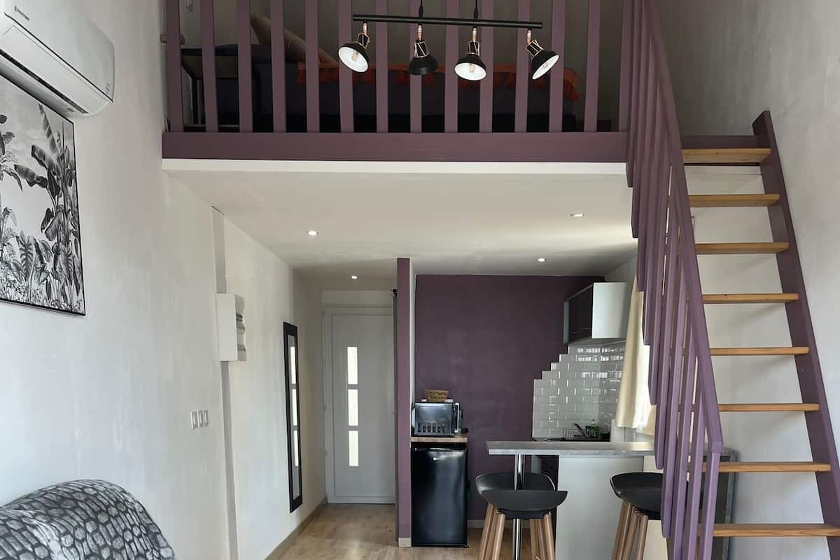 A modern living room with a blue sofa, a small kitchen area, and a wooden staircase.
