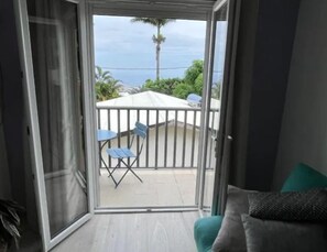 Una habitación con puerta corrediza de vidrio que da a un balcón con una sombrilla blanca, dos sillas azules y vistas al océano y palmeras.