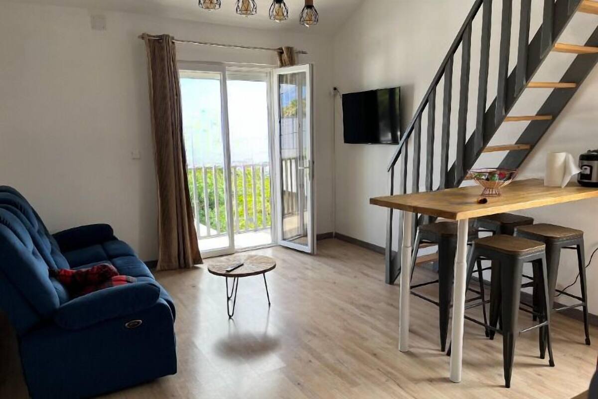 A modern living room with a blue sofa, a wooden dining table with stools, a staircase, and a sliding glass door leading to a balcony.