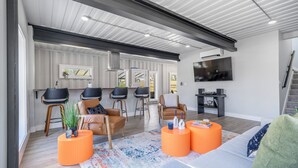 A modern living room with a sofa, two armchairs, a coffee table, and a TV. There are two bar stools and a kitchen area visible in the background.