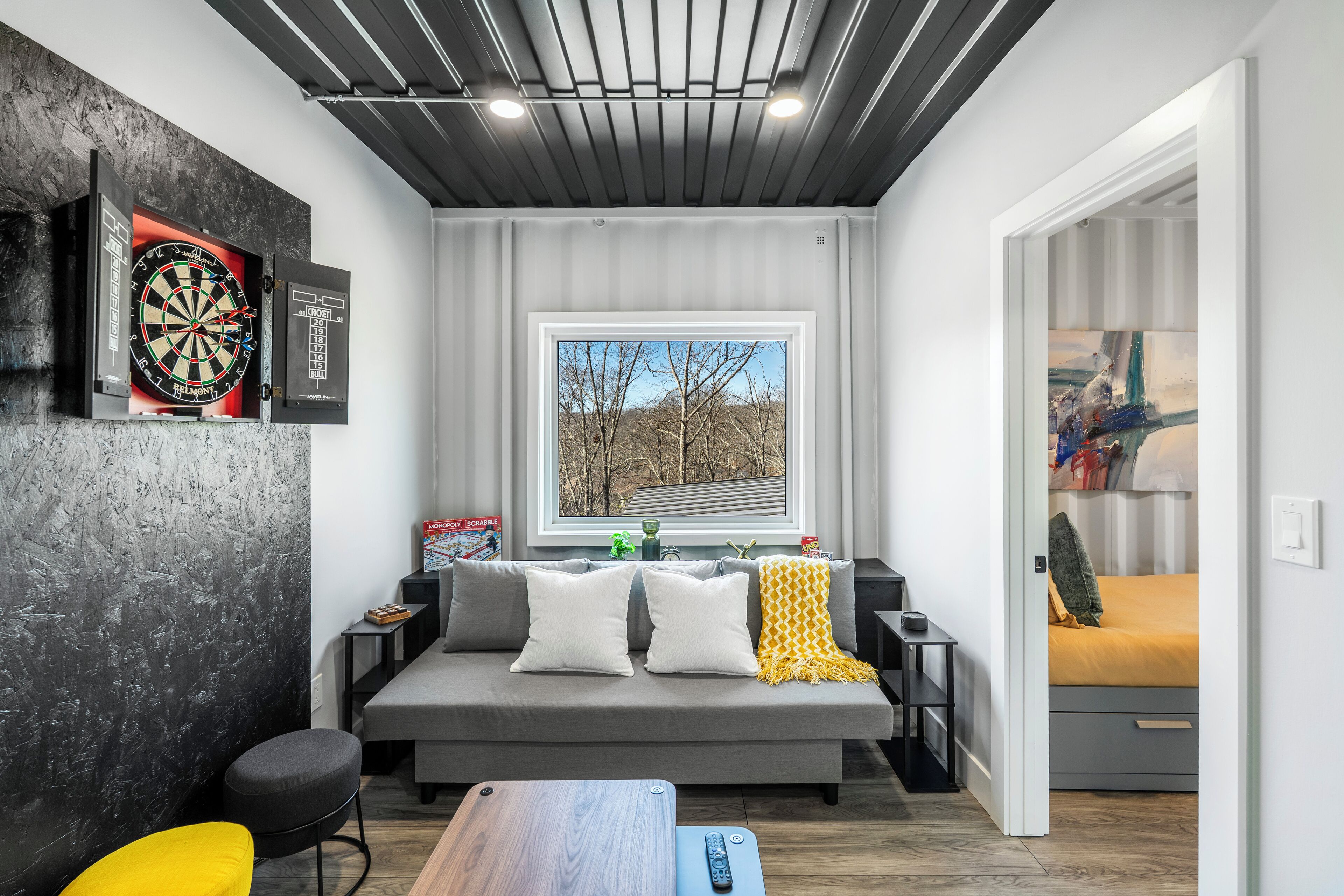 A modern living room with a grey sofa, a dartboard, and a window with a view of trees.