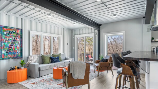 A modern living room with a grey sofa, orange ottoman, and a wooden bar stool.
