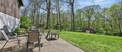 An outdoor patio with chairs and tables, a small shed, and a grassy area with trees.