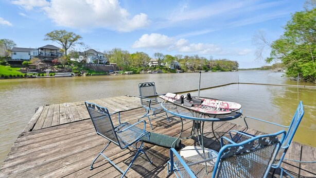 Un muelle de madera con sillas y una mesa de metal, con vista a un río tranquilo y casas al fondo.