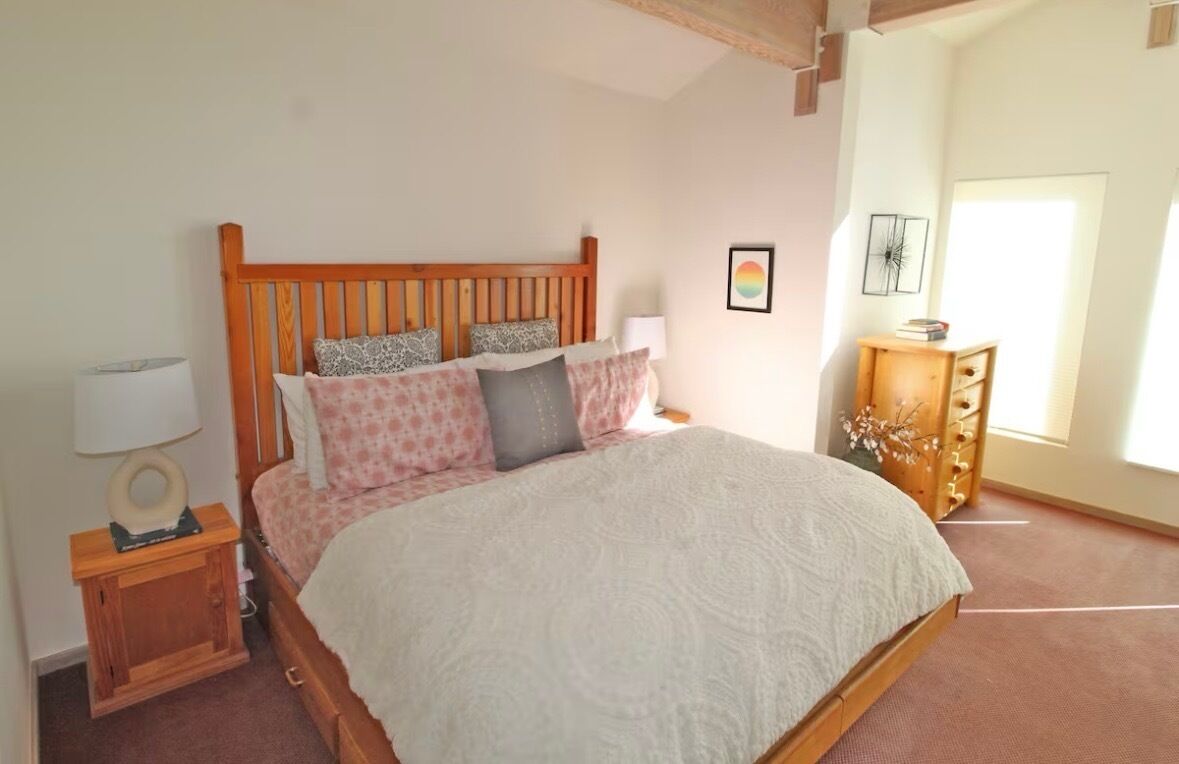 Une chambre à coucher avec un lit en bois, une table de chevet avec une lampe, une commode et une fenêtre laissant entrer la lumière naturelle.