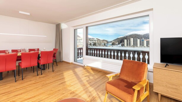 A modern room with a dining area, a large window with a view of a snowy mountain and buildings, a wooden floor, and a TV on a wooden cabinet.