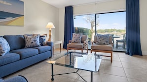 Ein Wohnzimmer mit einer Couch, Sesseln, einem Glastisch und Blick ins Freie durch eine Schiebetür.