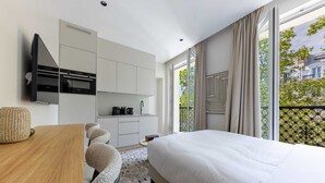 A modern hotel room with a large bed, a desk with a chair, a kitchenette, and a view of the city through large windows. - Marais - Studio in Paris (Paris)