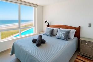 A bedroom with a large window overlooking a beach and a pool, a bed with a headboard, and a nightstand with a lamp.