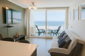 A room with a television, a dining table, a sofa, and a view of the ocean through a sliding glass door.