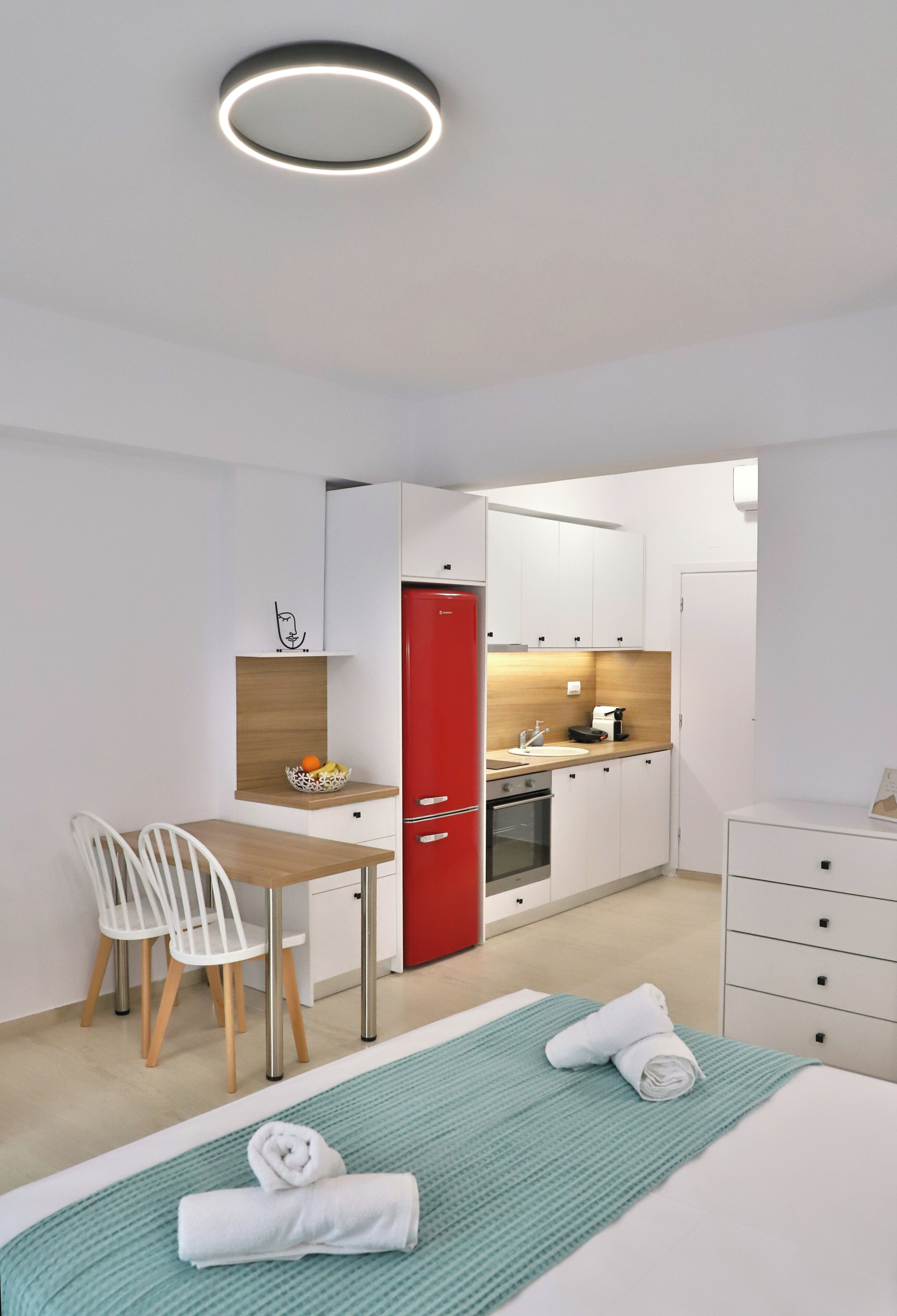 Una habitación de hotel moderna con una cama, un escritorio, un refrigerador rojo, un lavamanos y una pequeña cocina.