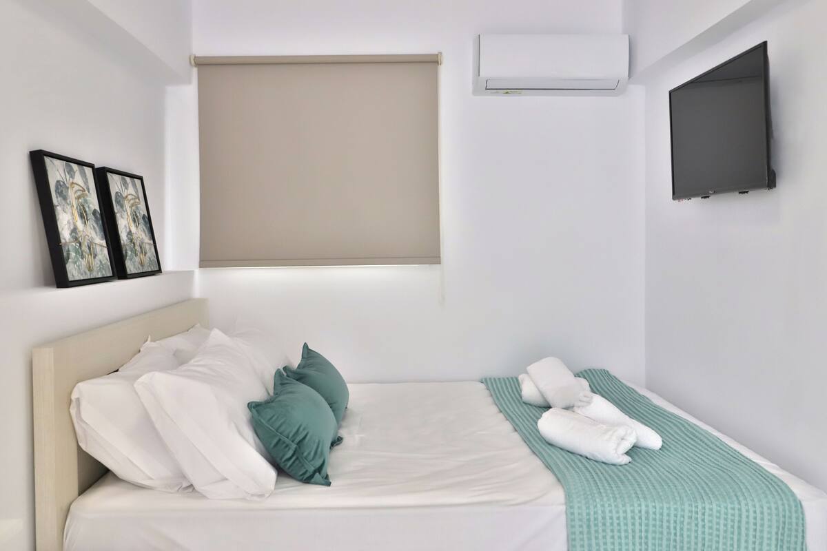 Un dormitorio con cama, almohadas, una colcha, un televisor montado en la pared y un equipo de aire acondicionado.