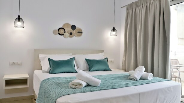 A neatly made bed with a teal throw blanket and two pillows, a wall-mounted light fixture, and a window with curtains. - Centro Apartments By Comfortbnb (Sitia)