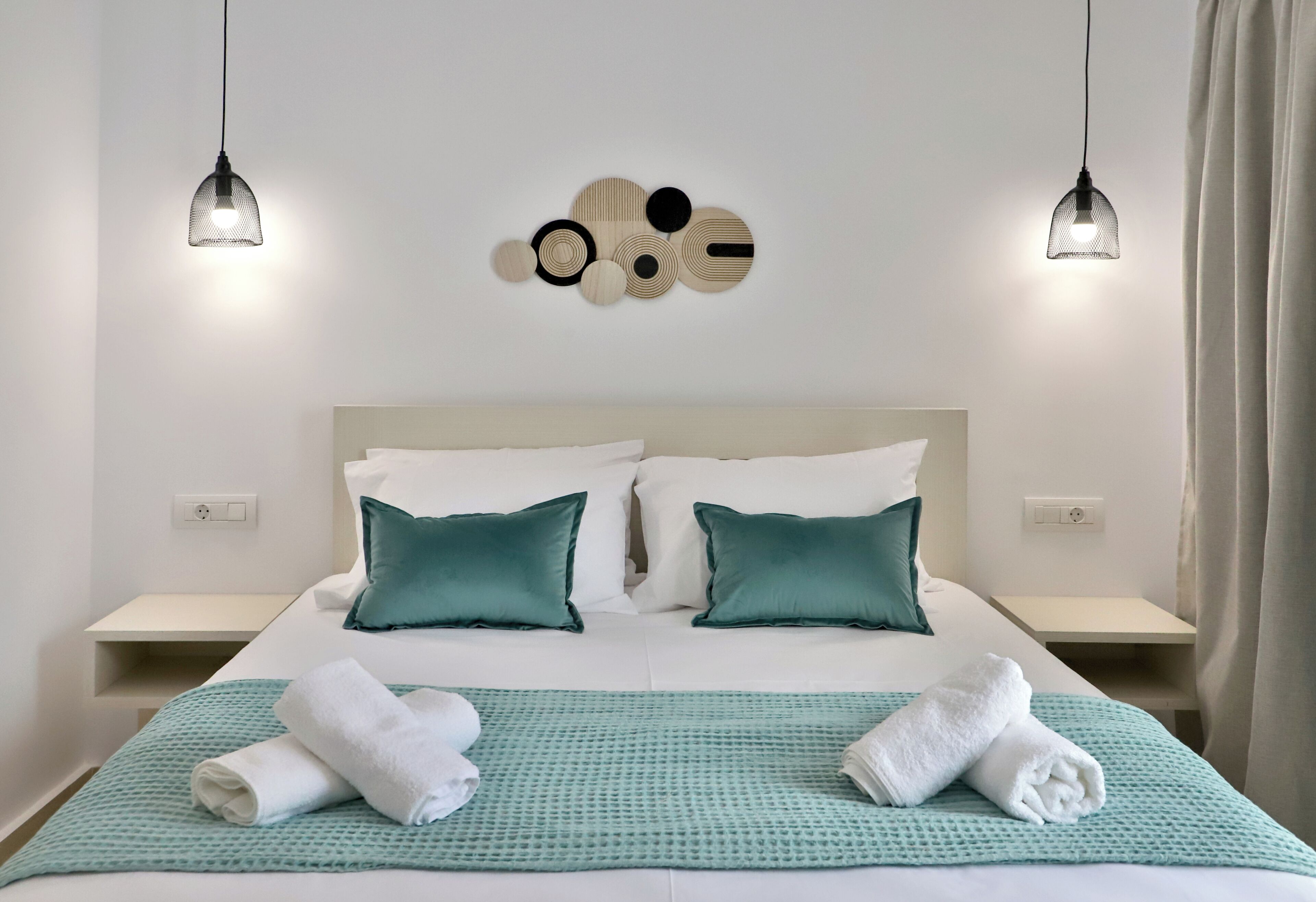 A hotel room with a bed, two bedside tables, a wall art piece, and two pendant lights.