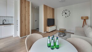 A modern living room with a white sofa, a round coffee table with bottles, a TV on a wooden cabinet, and a clock on the wall.