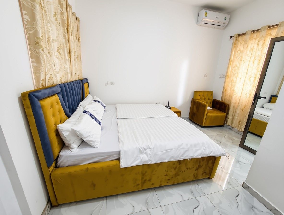 A neatly made bed with a headboard, a bedside table, and a chair in a room with a tiled floor and a door with a mirror.