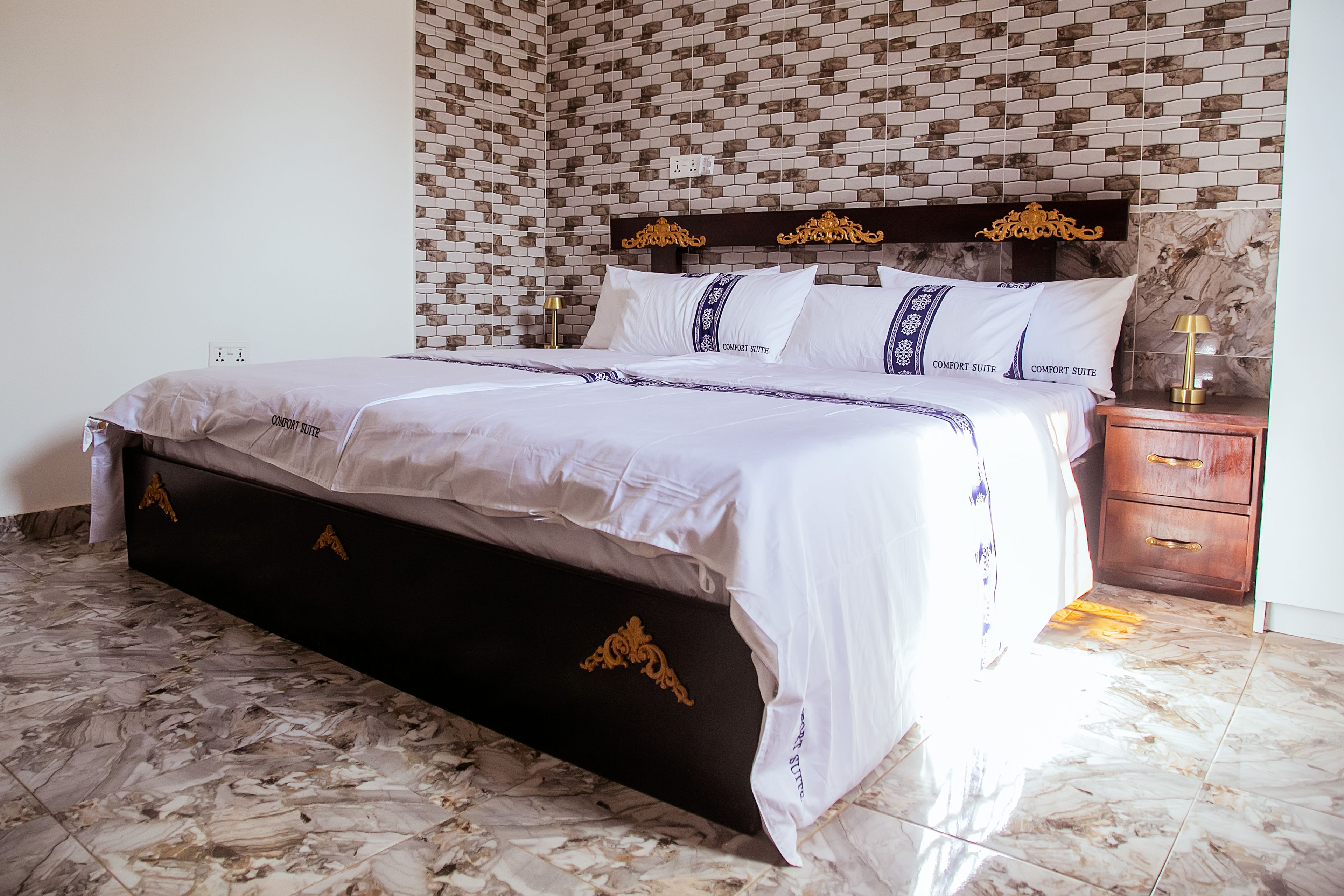 A bed with a dark headboard and gold accents, a bedside table with a lamp, and a patterned wall.
