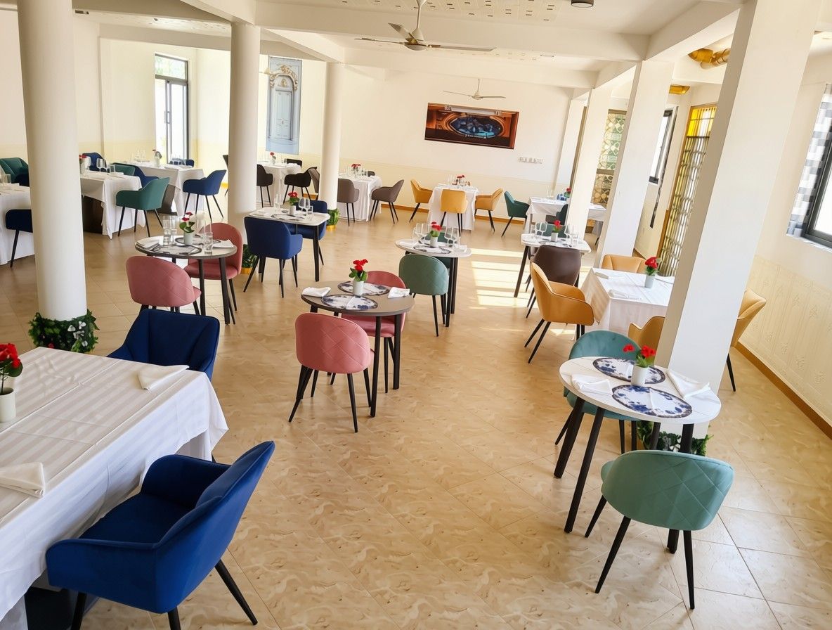 Ampla sala de jantar com várias mesas e cadeiras, uma televisão na parede e grandes janelas que proporcionam muita luz natural.