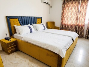 A double bed with a headboard, a bedside table with drawers, a chair, and a patterned curtain. - CK Suites (Adentan)