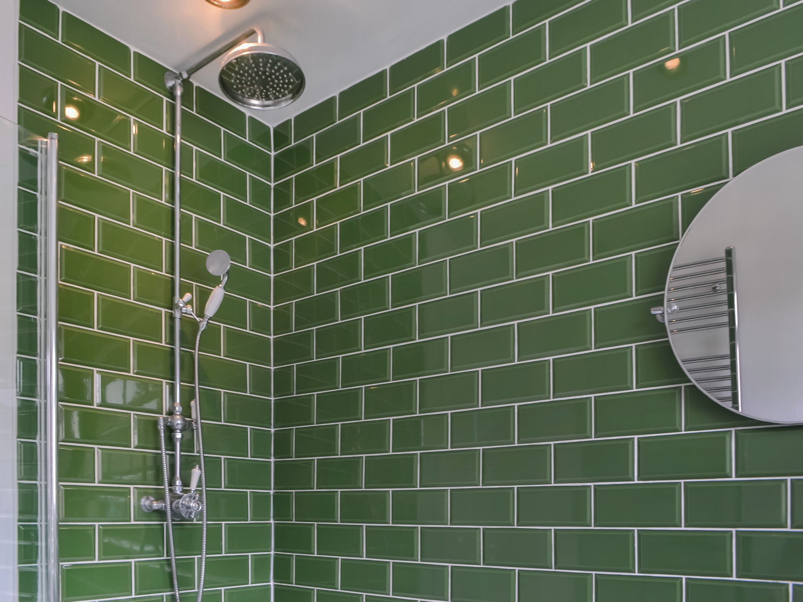 Une douche d’angle avec des carreaux verts, un miroir rond et un pommeau de douche.