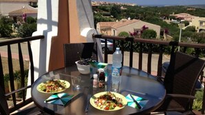 A round glass table with a meal, a bottle of water, and condiments on a balcony overlooking a coastal town. - Delightful sea View Apartment With Veranda (Stintino)