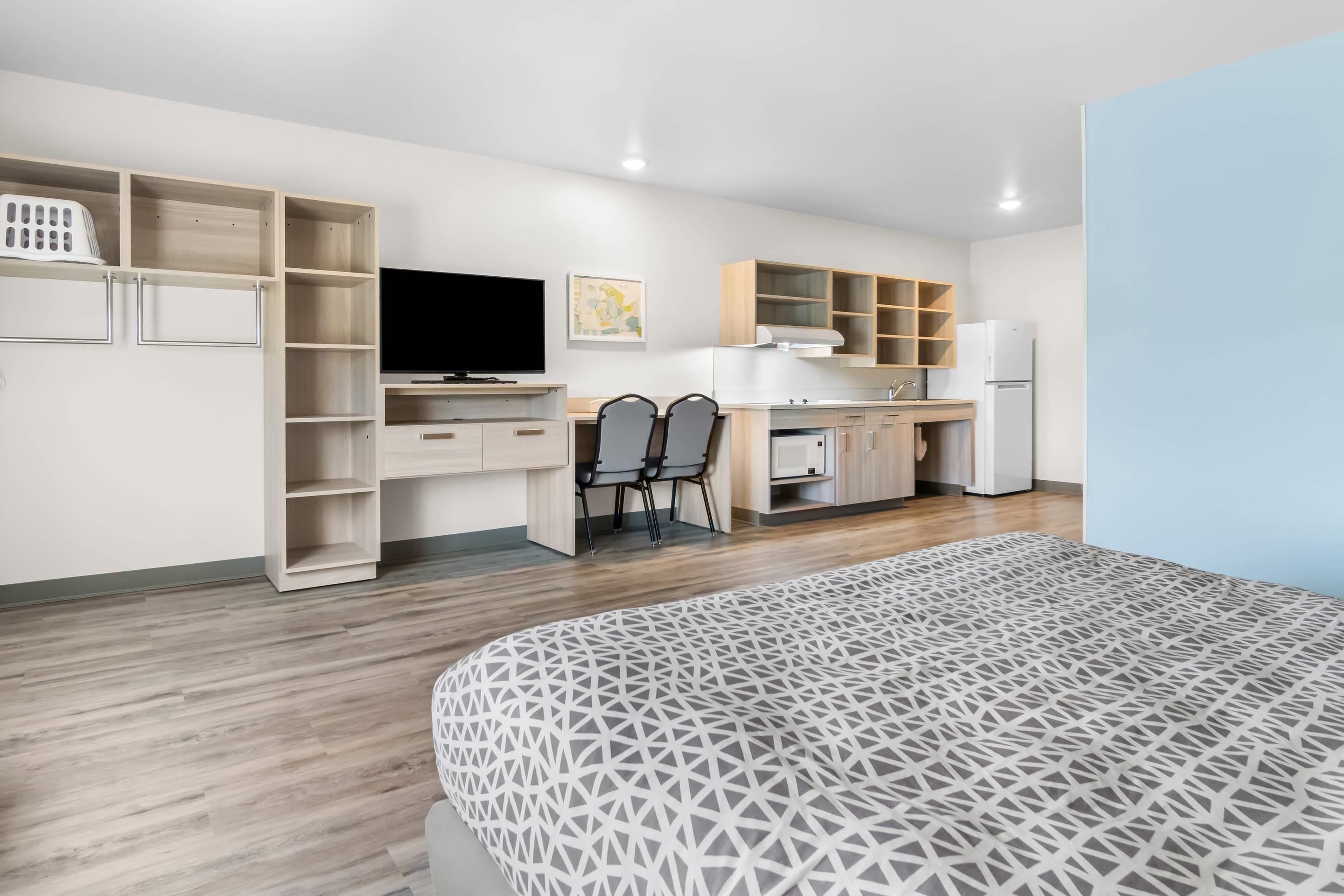 A modern hotel room with a bed, a desk, a chair, a television, and a kitchenette.