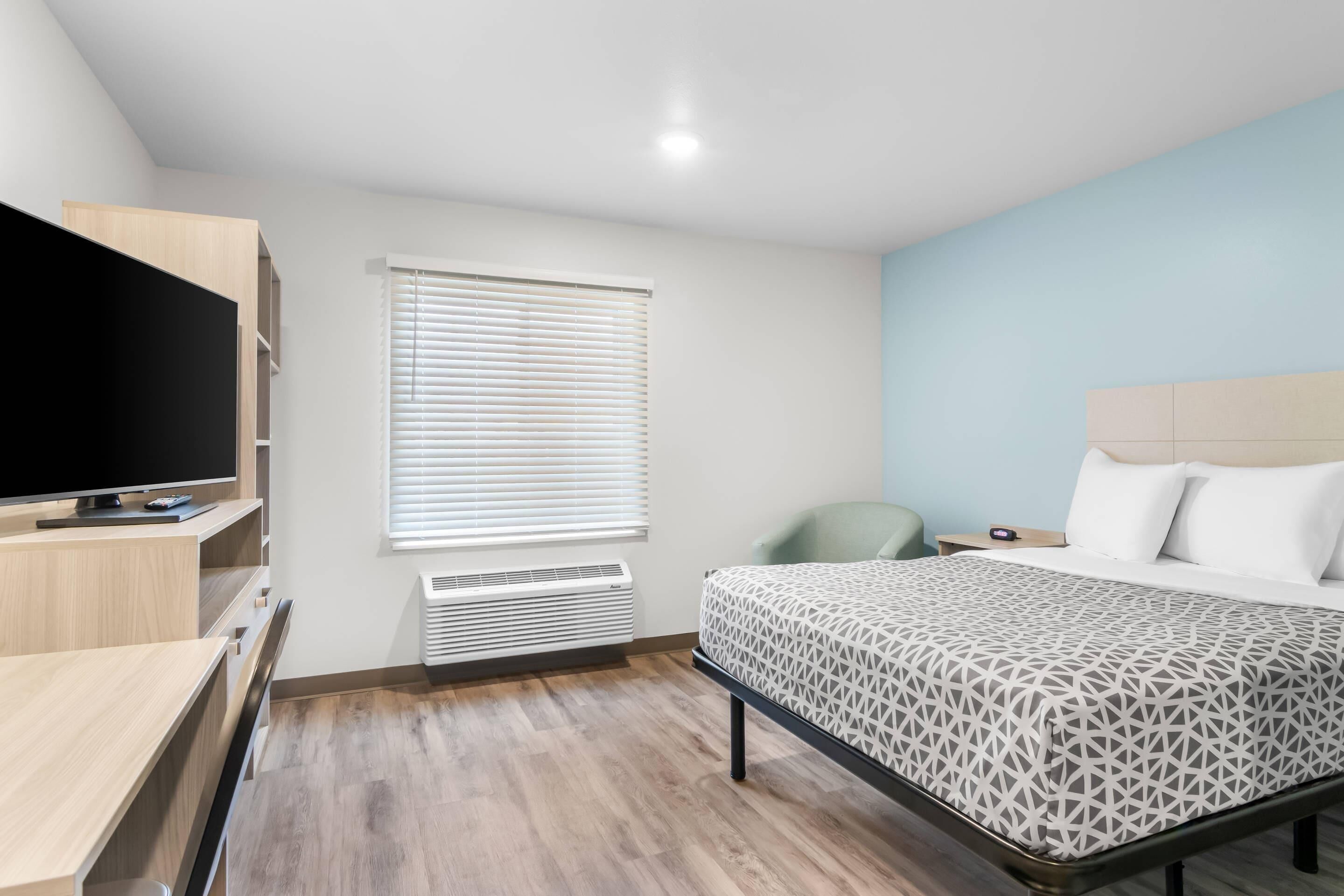 A modern hotel room with a bed, a desk, a TV, and a window with blinds.
