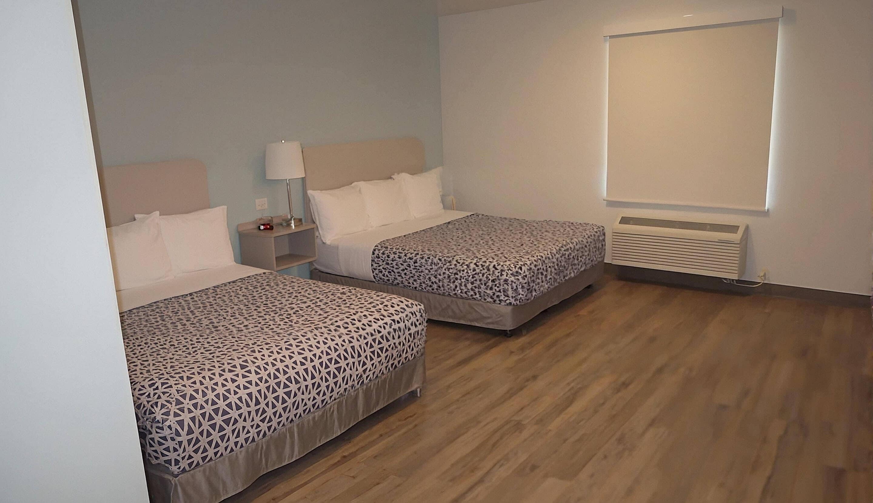 Habitación de hotel con dos camas, una mesita de noche con lámpara, una ventana con persianas y un aire acondicionado instalado en la pared.