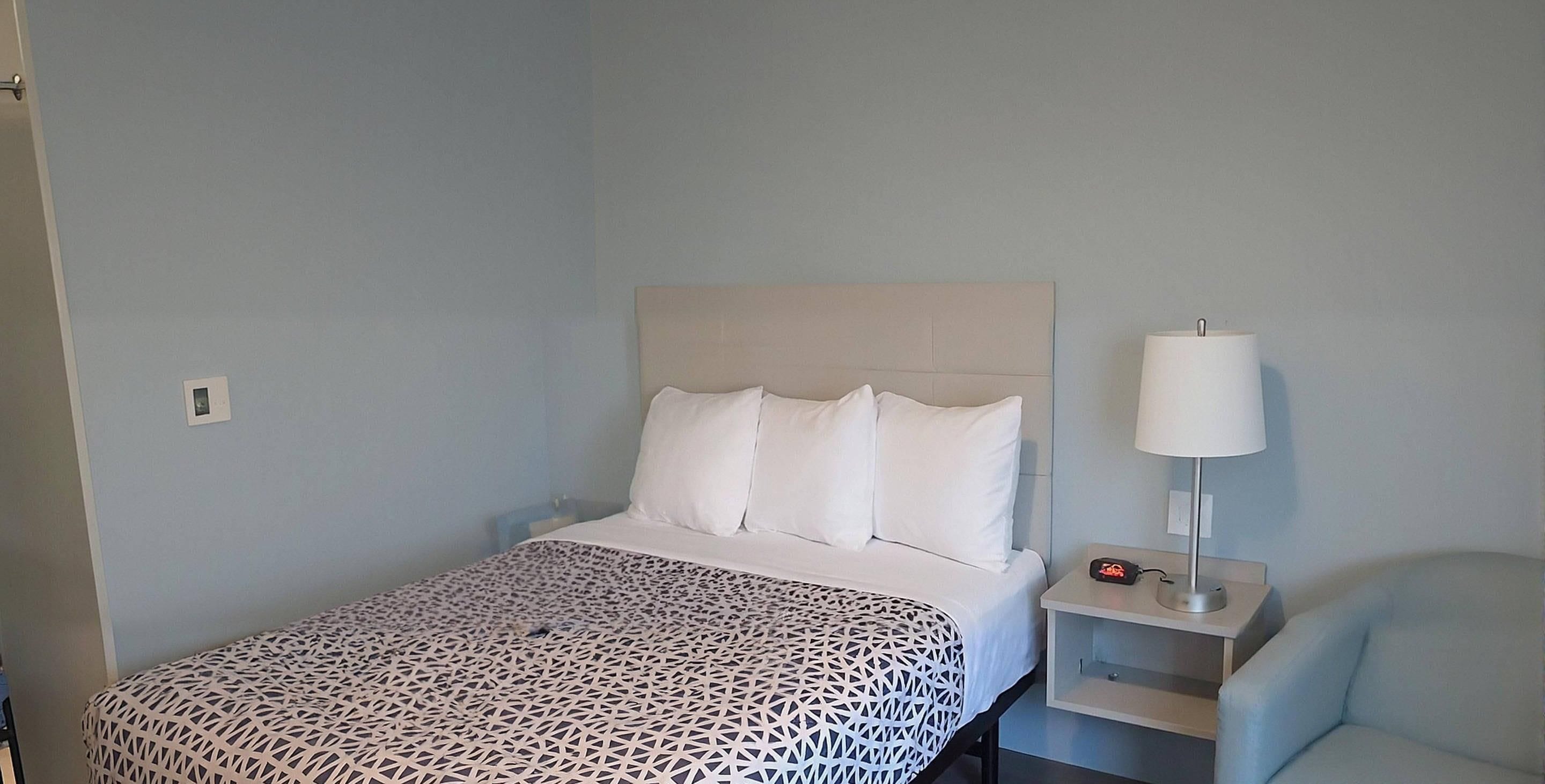 A neatly made bed with a patterned bedspread, a bedside table with a lamp, and a chair in a room with light blue walls.