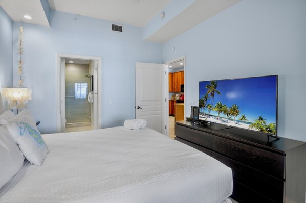 A hotel room with a bed, a TV displaying a beach scene, a bathroom with a shower, and a kitchen area visible through an open door.