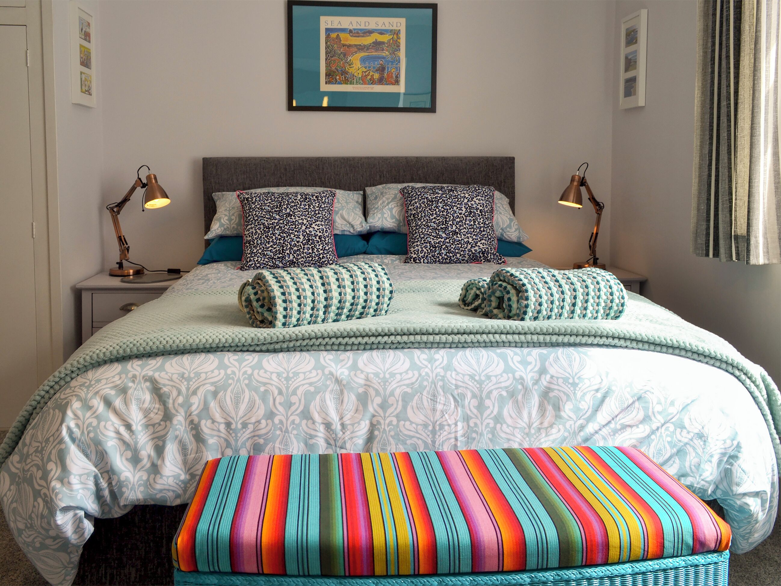 Uma cama bem arrumada com roupa de cama estampada e almofadas, um banco listrado colorido e um quadro na parede.