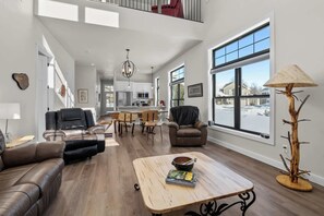 A spacious living area with a high ceiling, a dining area, and large windows offering views of the outside.
