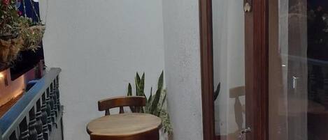 Un pequeño balcón con una mesa y sillas de madera, una planta en maceta y una puerta corrediza de vidrio.