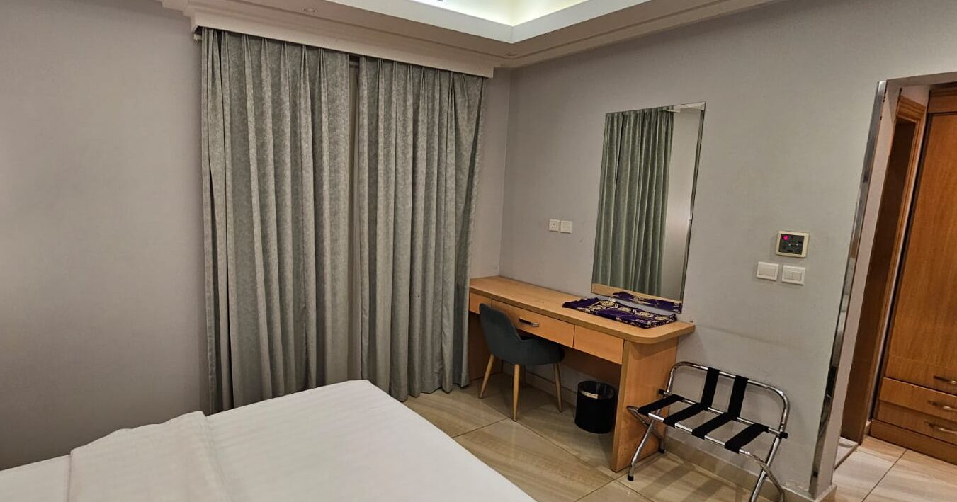 Quarto de hotel com cama, escrivaninha, cadeira, espelho e porta.