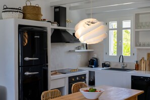 A modern kitchen with a black refrigerator, a built-in oven, a stove, and a sink. There is a dining table with a bowl of fruit and a hanging light fixture above it.