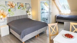 Un dormitorio con cama, mesitas de noche, una pequeña mesa con fresas, una silla, un baño con puerta de vidrio y un tragaluz.