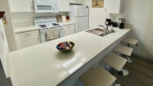 A modern kitchen with white cabinets, a microwave, stove, refrigerator, and a long countertop with stools. - 2-Bedroom Condo - close to all! (San Diego)