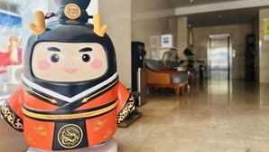 Une figurine décorative à la tête ronde, vêtue d’une tenue rouge et noire, placée dans une pièce dont le sol est recouvert de carreaux et où l’on aperçoit un bureau au fond.