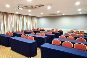 Sala de conferências com mesas e cadeiras dispostas em fileiras, um projetor e uma tela grande.