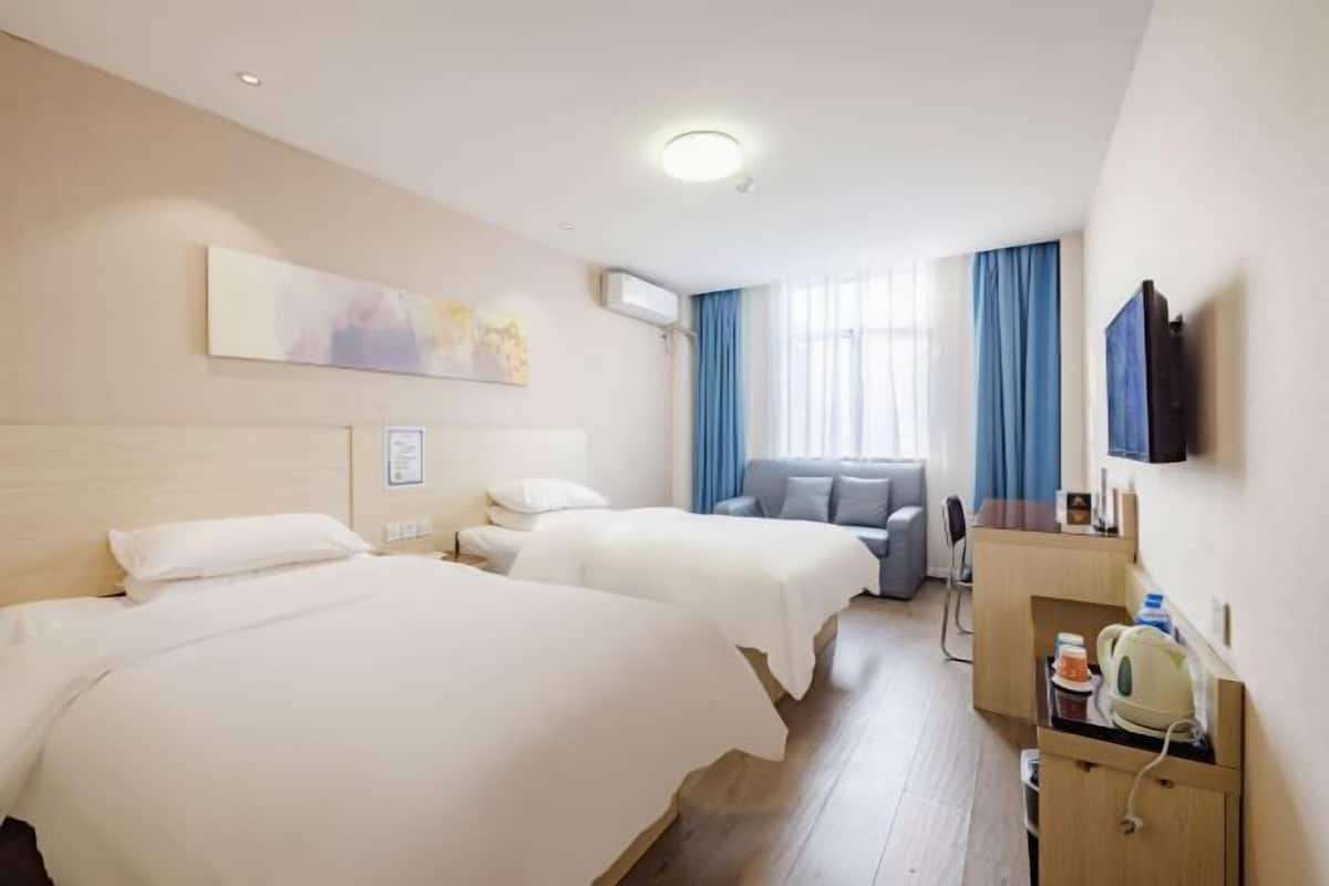 Quarto de hotel com duas camas, uma escrivaninha com chaleira, TV de tela plana e uma janela com cortinas azuis.