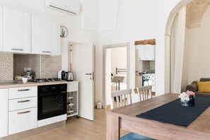 Cocina moderna con armarios blancos, estufa a gas y horno empotrado. Cuenta con una zona de comedor con mesa y sillas, y una sala de estar con sofá y mesa de centro.