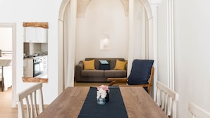 Un'area pranzo con un tavolo in legno, sedie e un vaso di fiori. Sullo sfondo, un divano con dei cuscini e si intravede una cucina attraverso un arco.