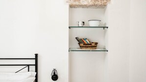 Una stanza minimalista con un letto, uno scaffale con libri, un bollitore e un cesto.