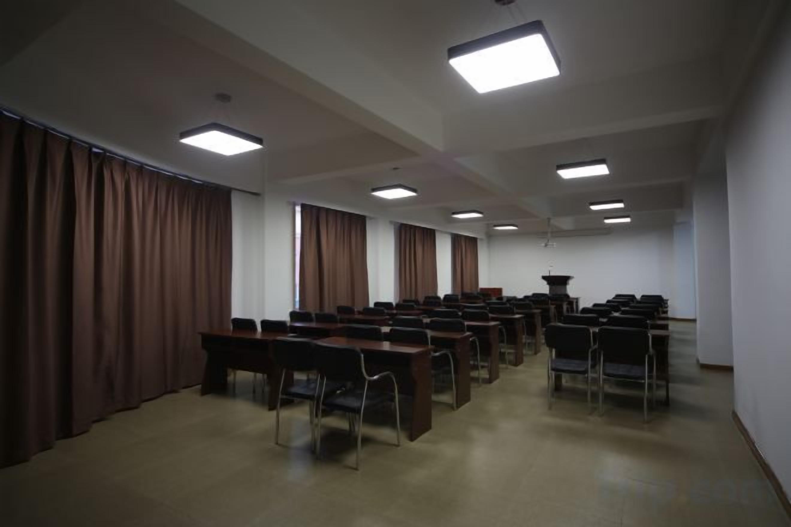 Un salón de conferencias con filas de mesas y sillas, ventanas amplias con cortinas e iluminación en el techo.