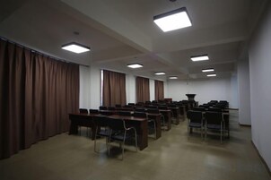 Un salón de conferencias con filas de mesas y sillas, ventanas amplias con cortinas e iluminación en el techo.