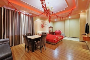 Une chambre d’hôtel avec une table à manger, des chaises, un lit avec une literie rouge, une télévision et un lustre.