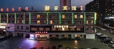 Un hotel con varios pisos, un estacionamiento y un edificio alto al fondo.